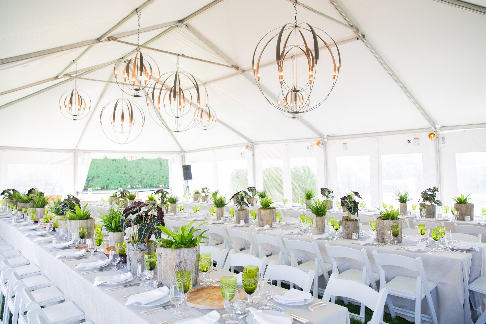 A white event tent in the Hamptons set for a dinner produced by Golden Circle's principal, with spherical chandeliers, long tables, and green glassware