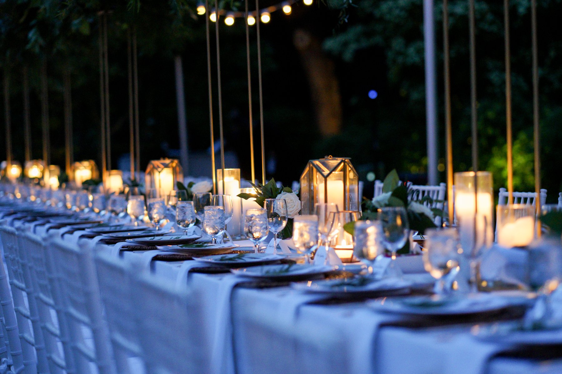 An outdoor Hamptons dinner table at dusk, produced by Golden Circle's principal, lit by candles and hurricane lanterns with white roses and string lights overhead