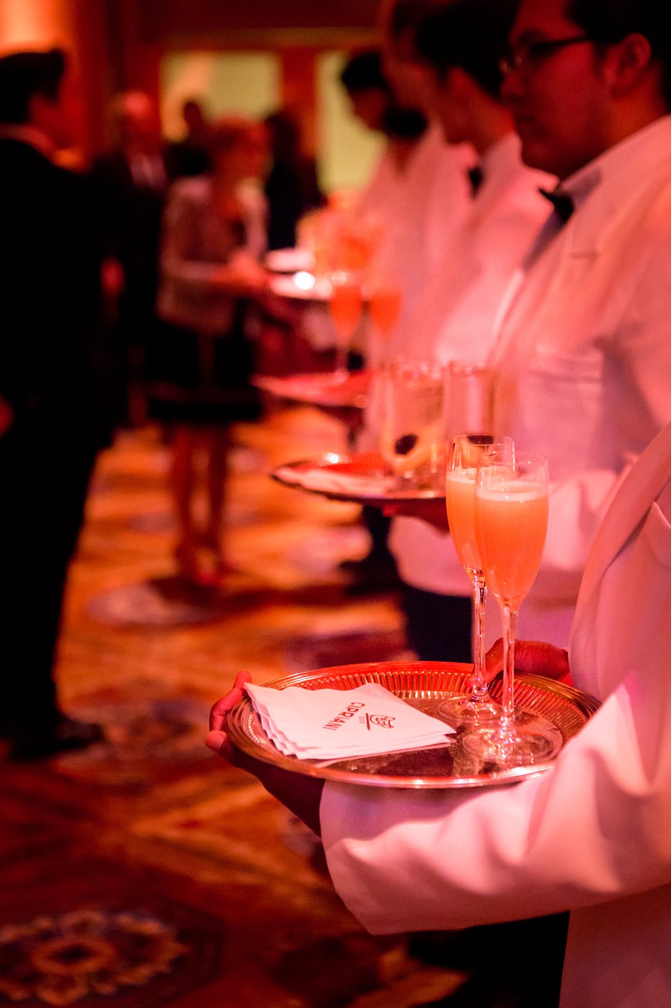 White-jacketed servers passing trays of champagne cocktails in a warm, red-lit ballroom at a New York City gala produced by Golden Circle's principal